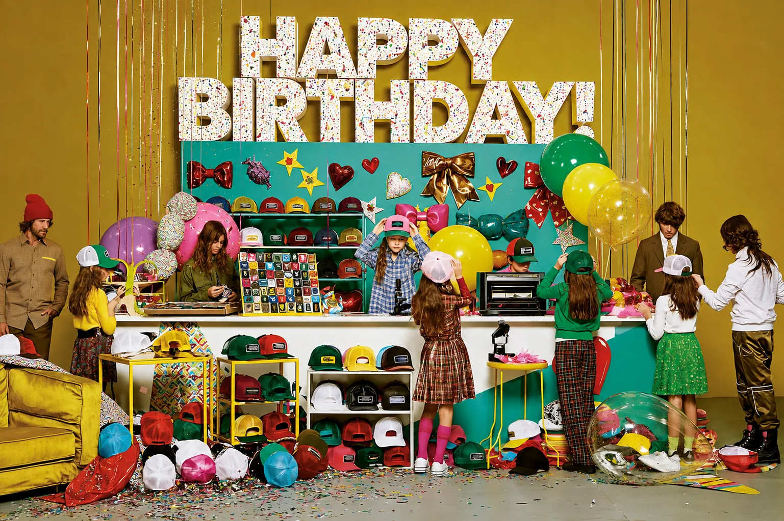 Maximalist Happy Birthday hat bar set by Brimstreet with giant confetti letters, shelves of colorful trucker hats, a patch board, and kids picking their birthday party hats
