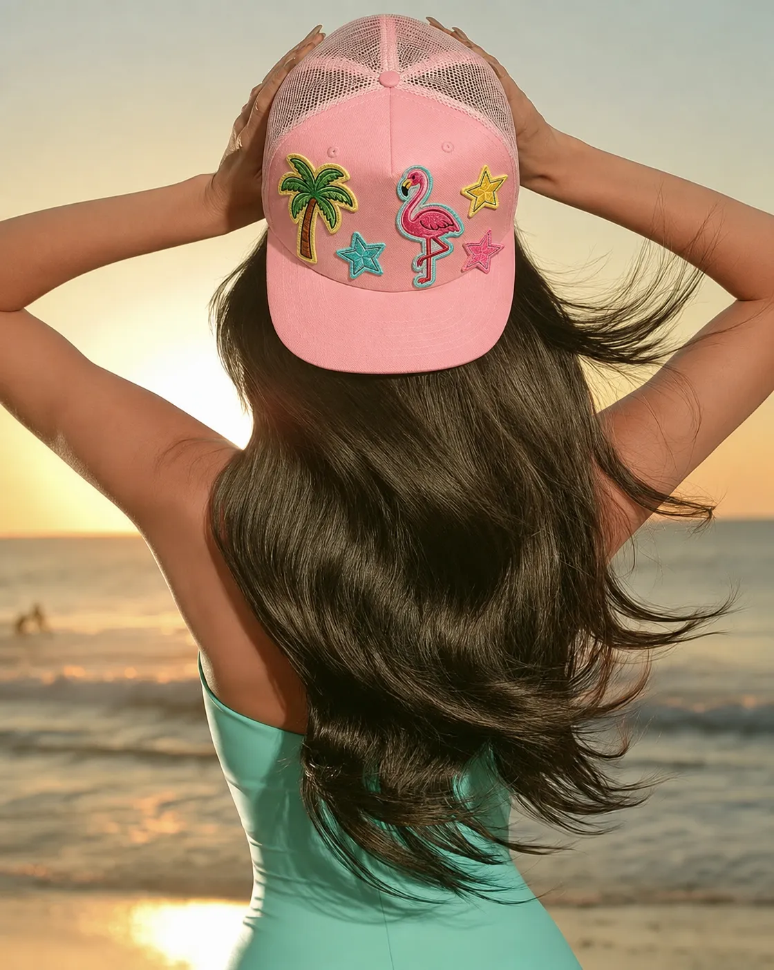 Woman on the beach at sunset wearing a pink custom trucker hat with palm tree, flamingo, and star patches from the Brimstreet hat bar
