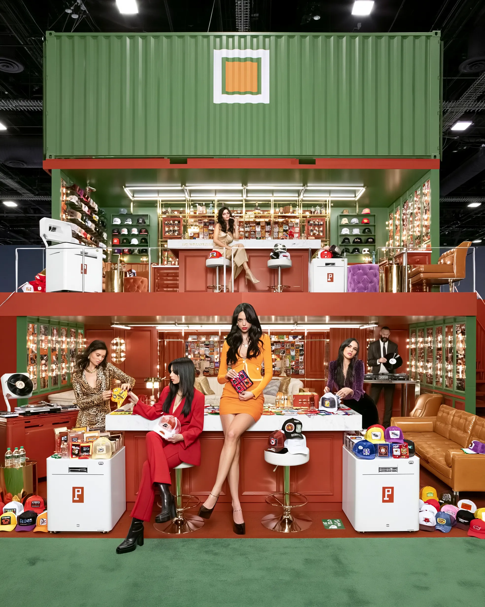 Two-story Brimstreet trade show booth built from shipping containers, with walls of custom trucker hats, patch-covered counters, lounge seating, and a DJ on the convention floor